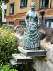Small bronze sculpture of Amelia Edwards at the top of steps formed by books projecting from the side of a stone wall.