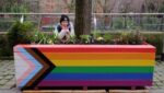 A smiling young woman (Qiuyan Chen) with face cupped in hands and elbows resting on a flower bed painted with trans and LGB colours