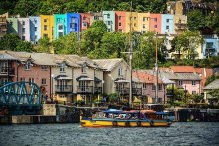 Small bright yellow and blue ferry boat sailing along Bristol harbour with multi-coloured terraced houses in the background