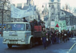 Demonstrators carrying banners with "National Gay News Defence Committee" walk behind a flat-top lorry in a city street with a band playing steel drums made from 55-gallon oil barrels
