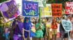 Six smiling young people wearing brightly coloured clothes are holding large signs with slogans such as "love wins" and "stand proud". Behind them is a large crowd about to start a street parade.
