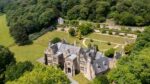 Aerial view looking down on a grand three storey stone manor house with an arched front door, steeply sloping roofs and tall chimneys, surrounded by sloping lawns bordered by stone walls and trees.