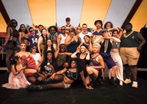 About 25 multi-ethnic flamboyantly-dressed young men, women and gender-queer people posing in a group, some standing, some squatting.