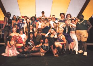 About 25 multi-race flamboyantly-dressed young men women and gender-queer people posing in a group, some standing, some squatting