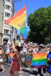 Colourfully dressed people waving Pride flags parade along a city street in bright sunshine.