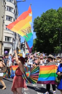 Colourfully dressed people waving Pride flags parade along a city street in bright sunshine.